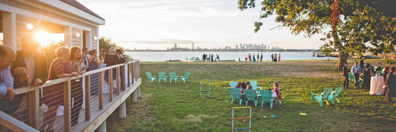 The image shows an outdoor gathering, likely a wedding or party, with people on a deck and on the grass. There are chairs set up, and a body of water is visible in the background. The lighting suggests it's either sunrise or sunset.